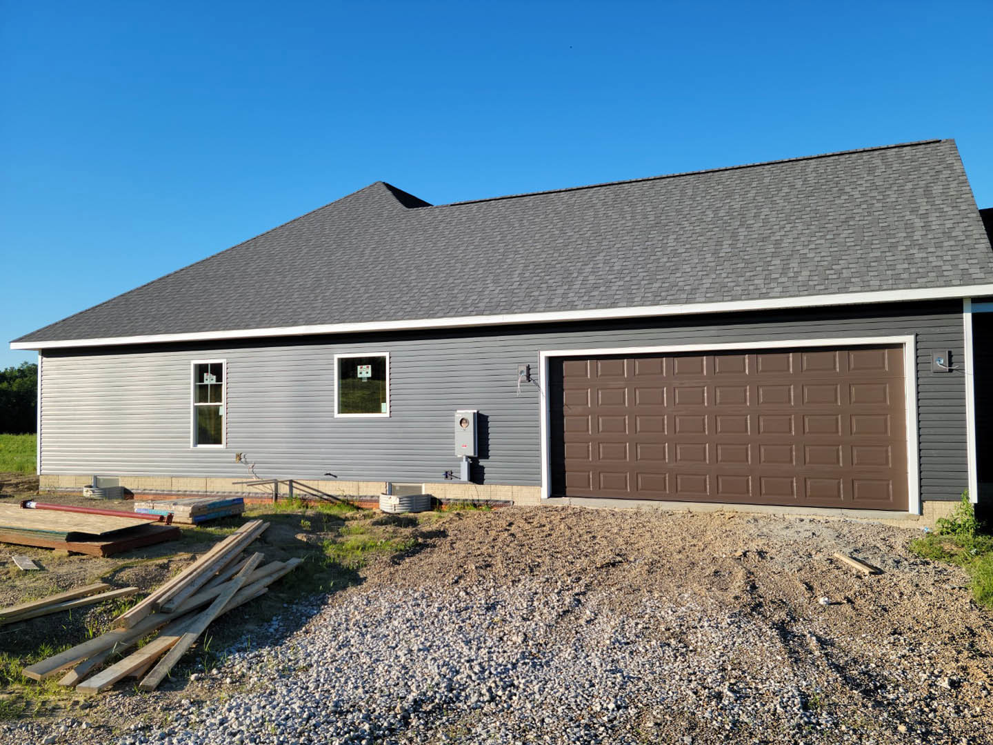 Brown garage door with white trim on a house, window displaying a sign, pile of wood and gravel ground in front, white siding and gable roof visible
