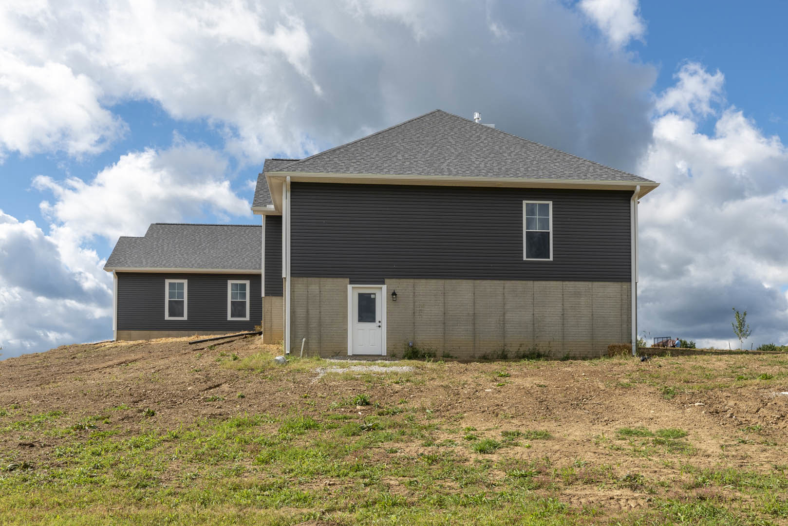 Gray exterior wall with white front door featuring a window, white-framed window beside entry, grassy and dirt yard in foreground, cloudy sky above