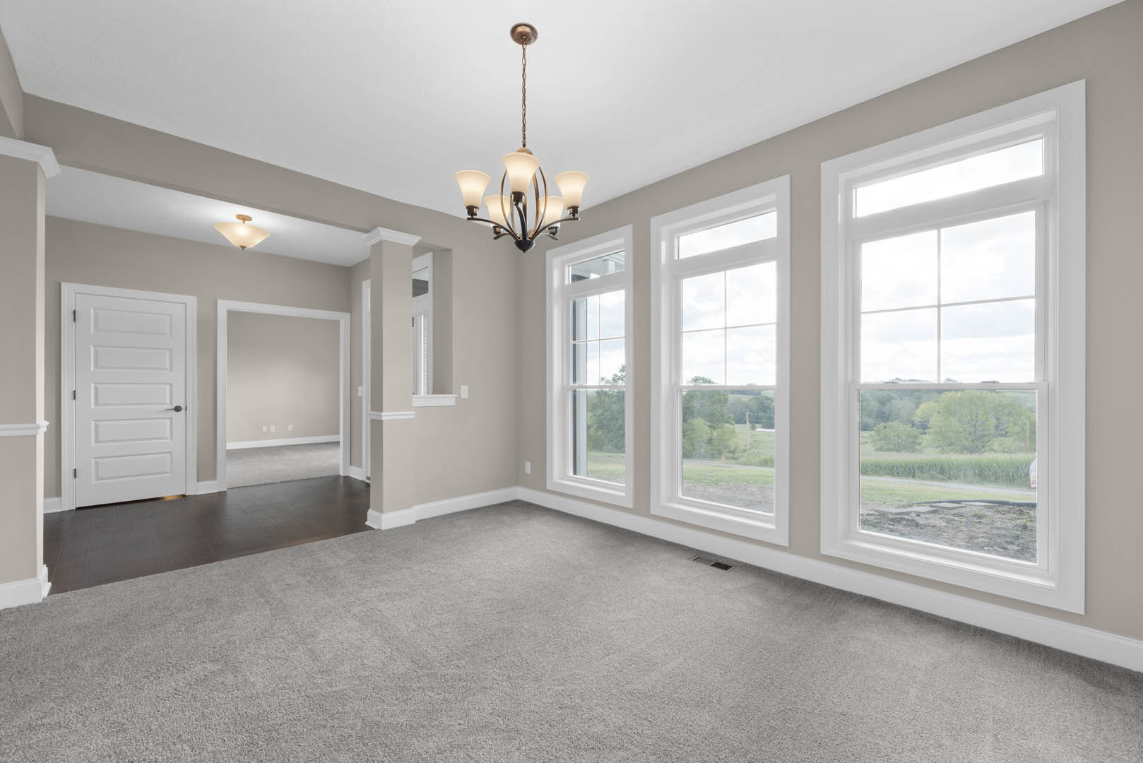 Carpeted room featuring a modern chandelier, large windows with white trim, white door with black handle, and views of trees and fields outside