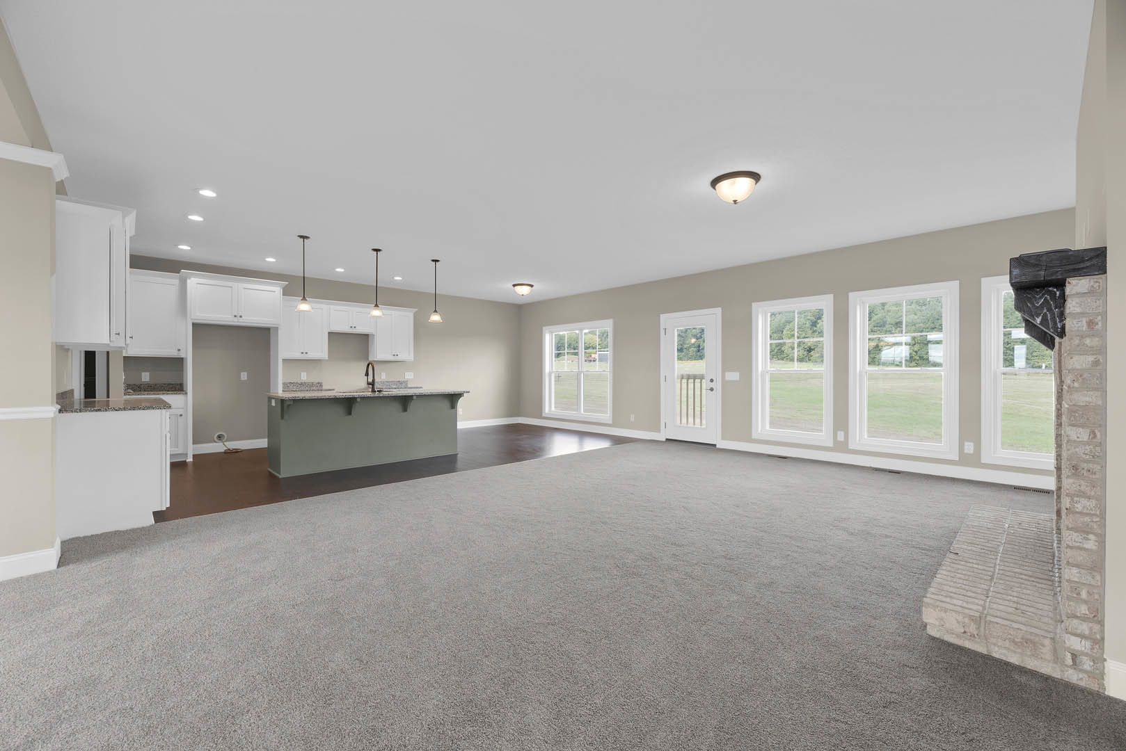 Spacious open floor plan featuring a modern kitchen with bar seating, grey accent wall, tile flooring, large windows overlooking a field of trees, and a white door with glass