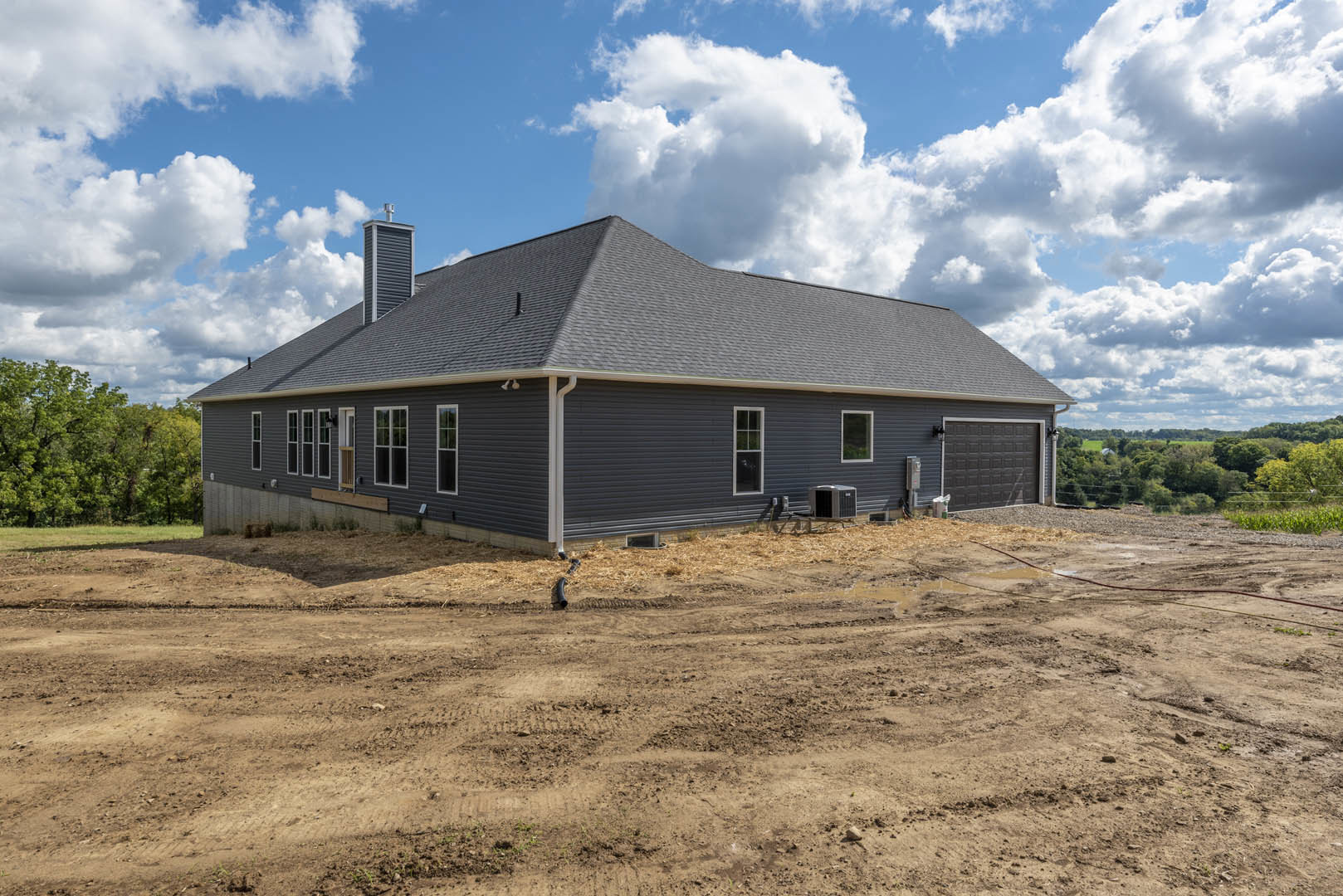 Framed house under construction with exposed roof, dirt field in foreground, scattered black pipe, and trees under partly cloudy blue sky