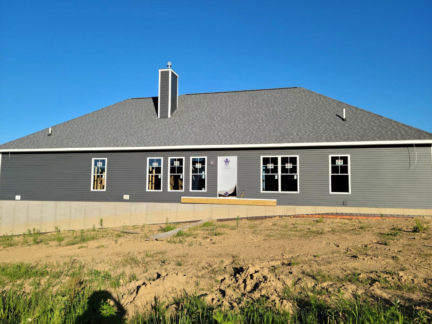 Grey house with white trim under construction, surrounded by dirt and sparse grass, blue sky overhead, white-framed window and fence visible, white utility box with sign in