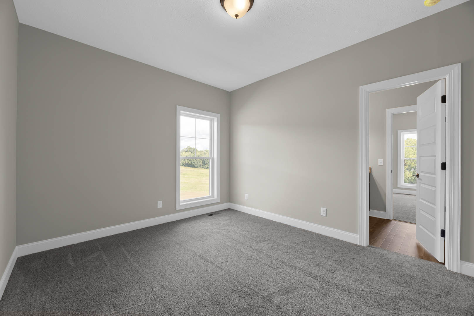Neutral-toned room with grey carpet, white walls, ceiling-mounted light fixture, white door, and window overlooking grassy field