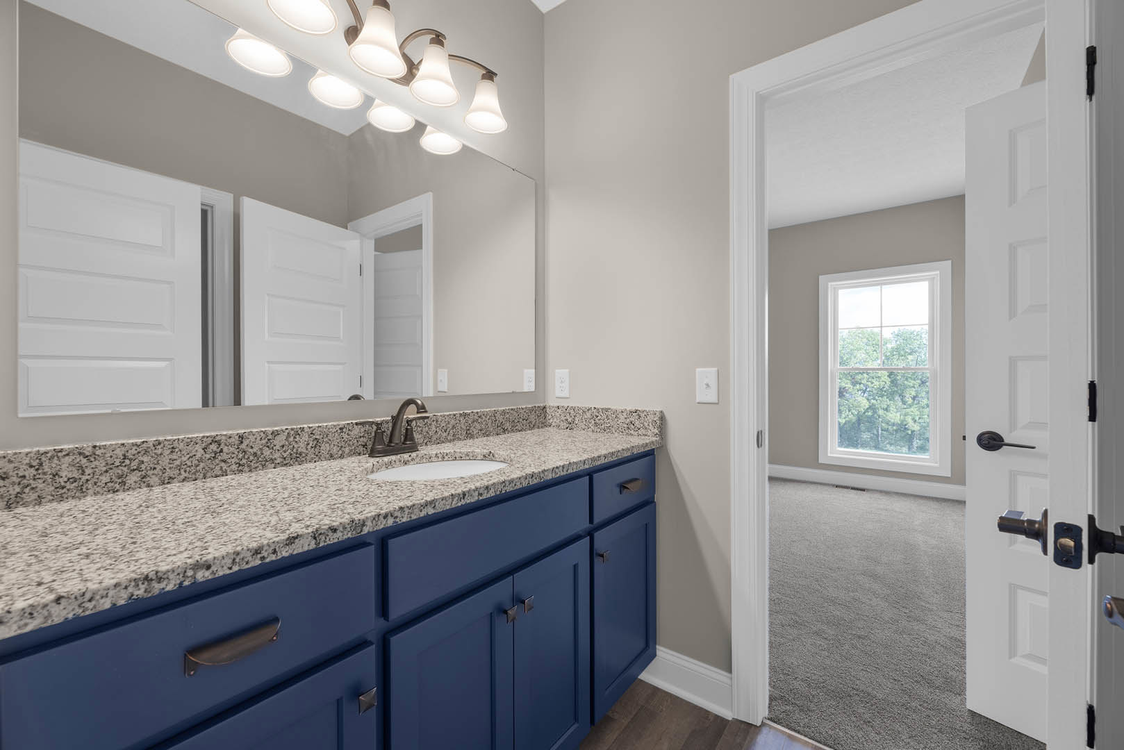 Bathroom with marble countertop, large mirror above blue vanity with sink and blue cabinets, white door open to hallway, window showing trees outside, modern light fixture on wall