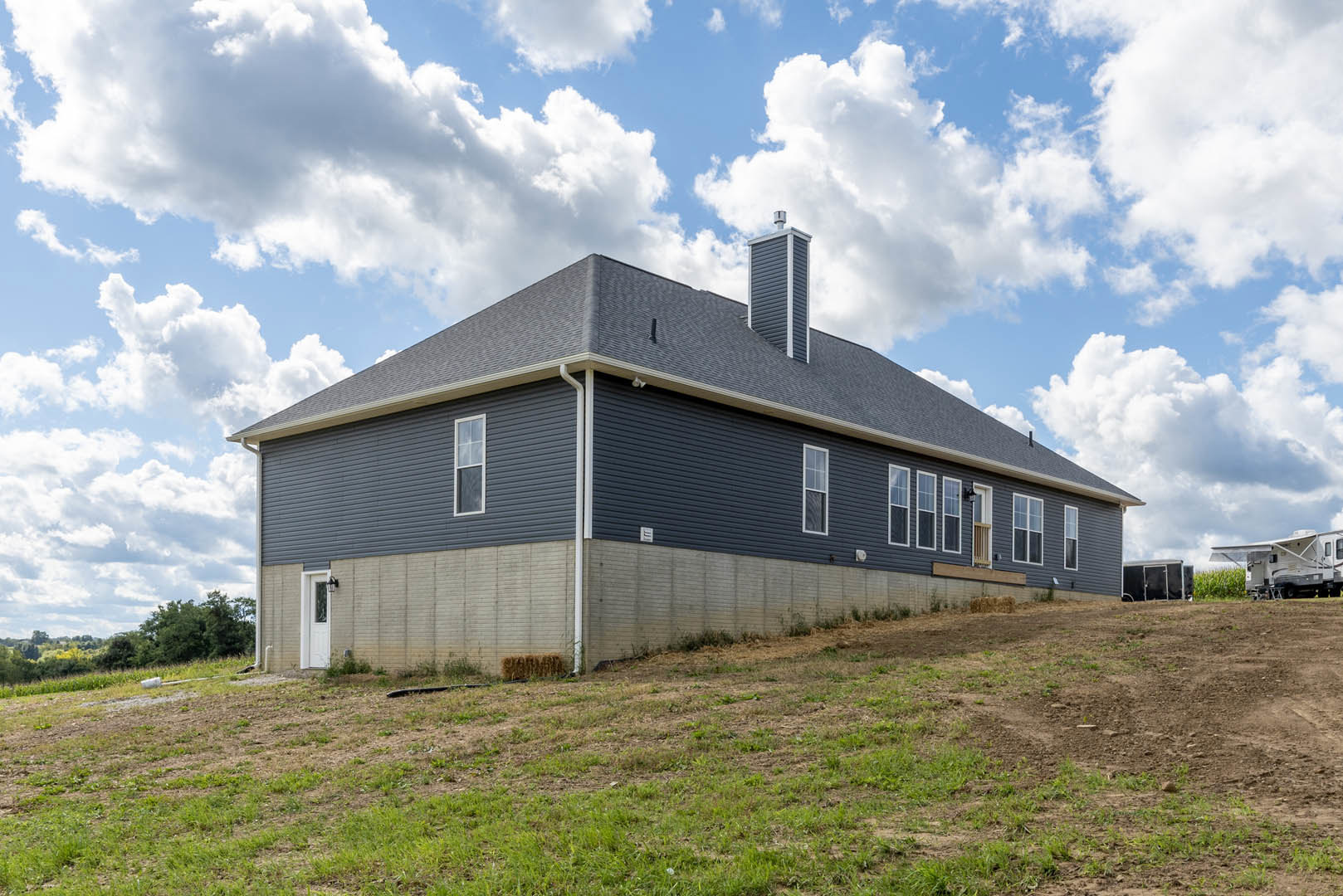 Two-story cottage with white siding and brick chimney, set atop a grassy hill under a cloudy sky, white RV parked beside the house
