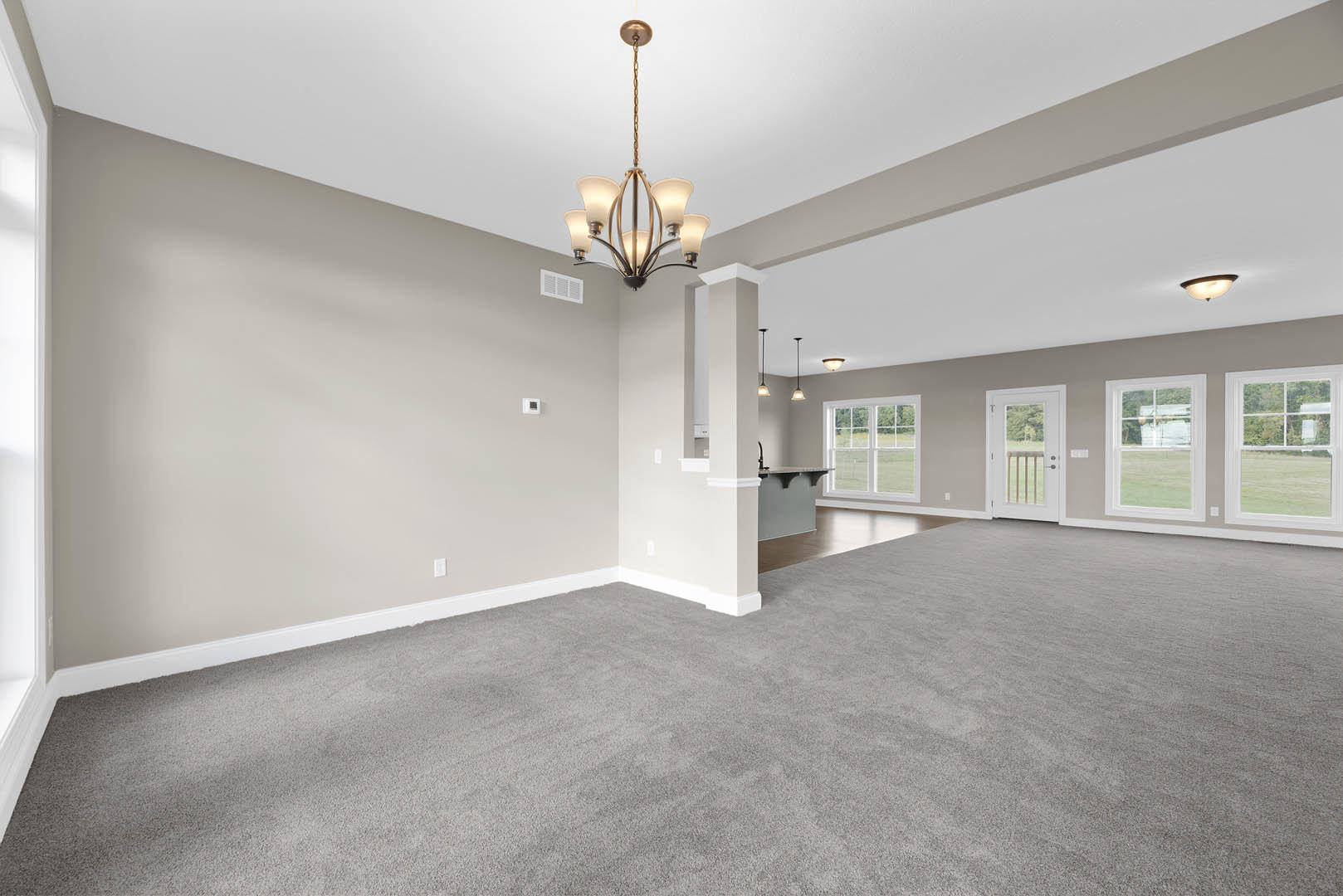 Open-concept living area with carpeted flooring, ornate chandelier, white kitchen cabinetry, glass-paneled door, and windows overlooking a green lawn