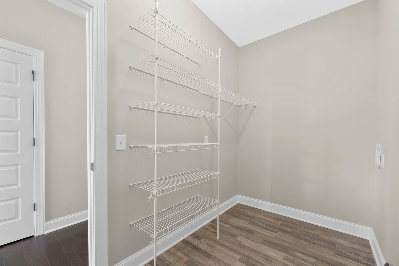 White wire shelving unit mounted on a white wall, close-up of a white door and light switch, wood floor with white baseboard trim.