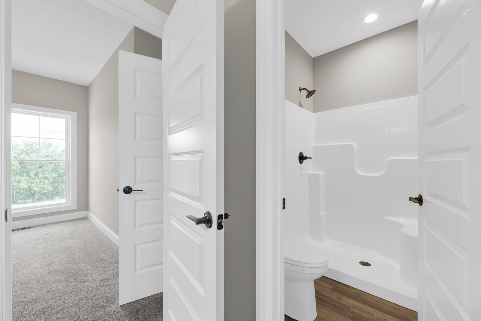 Bathroom with white paneled doors, glass shower enclosure, chrome door handle, white toilet, carpeted floor, window overlooking trees.