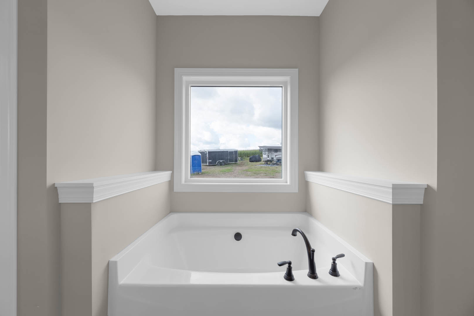 Freestanding bathtub beneath large window, white molding on wall, close-up of modern sink, view of field with black trailer outside