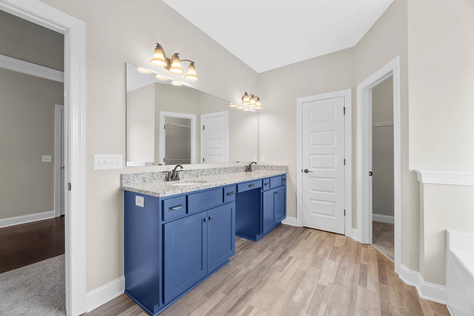 Bathroom with blue cabinetry, dual sinks set in a blue countertop, large mirror above, white door with black handle, white tile floor, modern light fixture illuminated, close-up of
