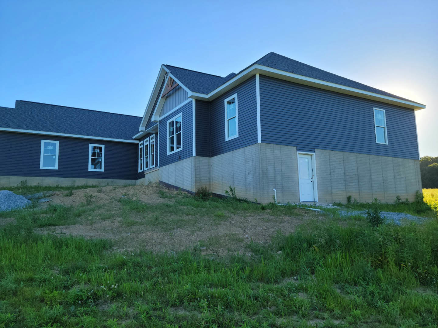 Two-story house with light-colored siding, white framed windows, and a white front door, set beside a grassy lawn and gentle hill under a clear sky.