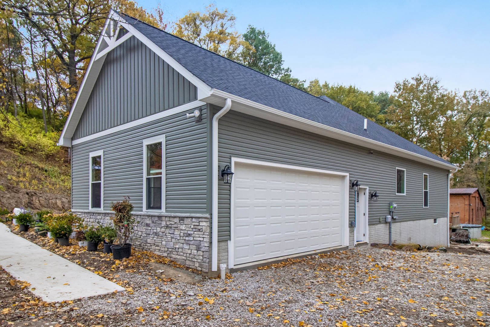 White garage door with lamp post beside brick house, white-framed window, sidewalk lined with potted plants, gravel and fallen leaves scattered on ground