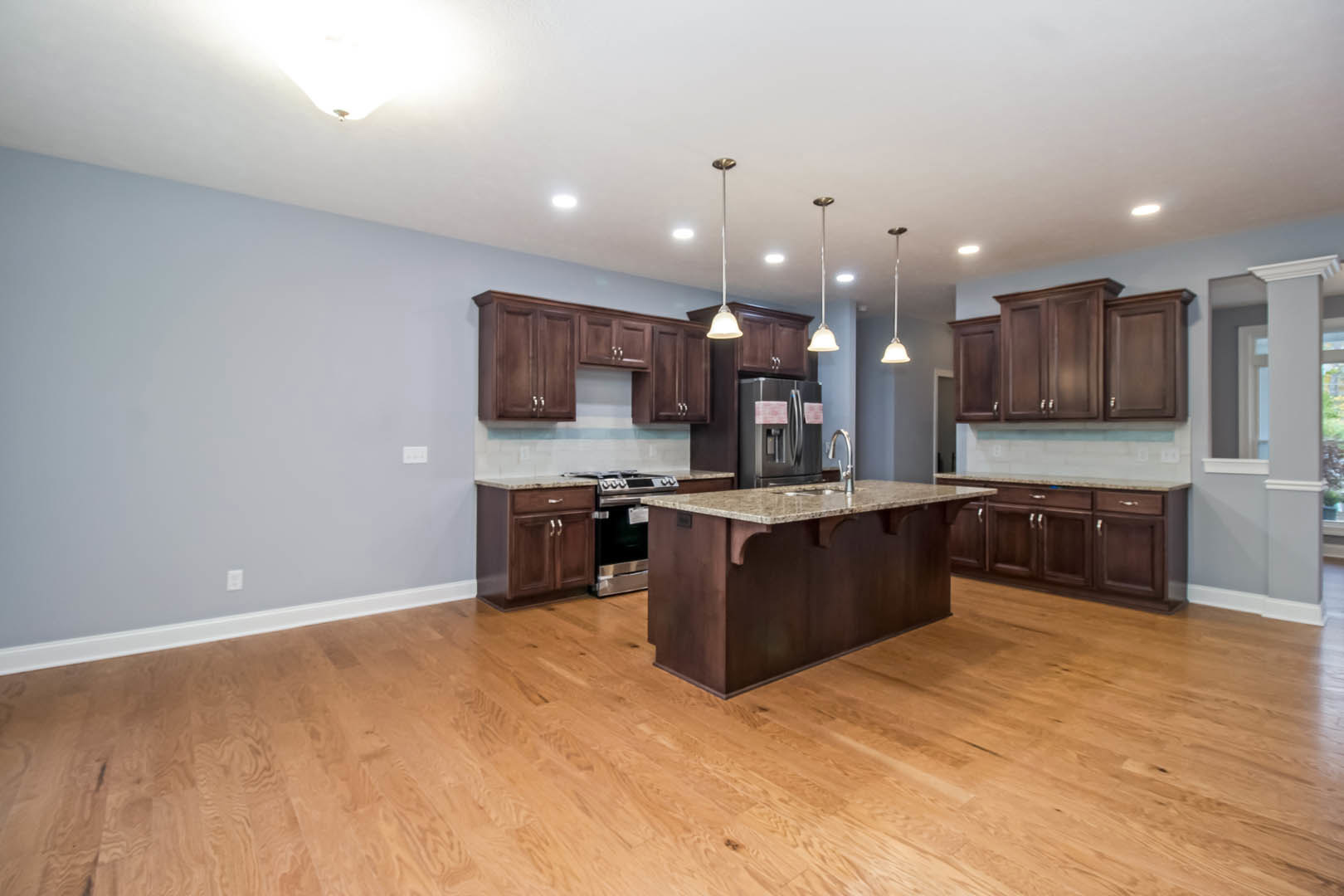 Spacious kitchen featuring a large central island with built-in sink, stainless steel stove, brown cabinetry, hardwood flooring, and stainless steel refrigerator.