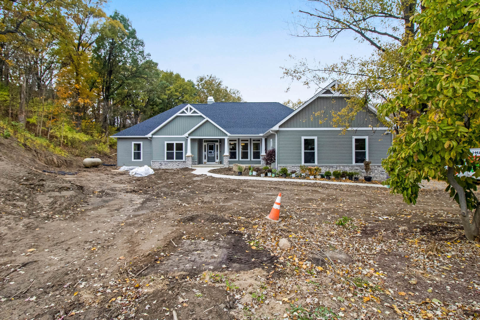 Two-story house with blue roof, white-framed windows, dirt driveway, and surrounding trees; orange traffic cone near construction area in foreground