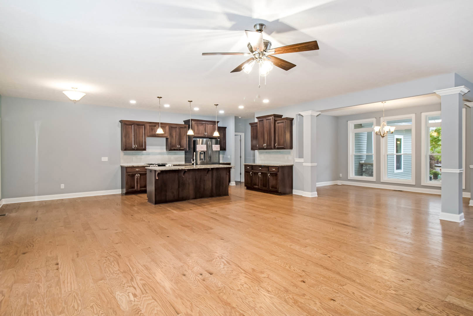 Spacious kitchen with hardwood floors, central island featuring drawers, stainless steel refrigerator, ceiling fan with lights, and white cabinetry.