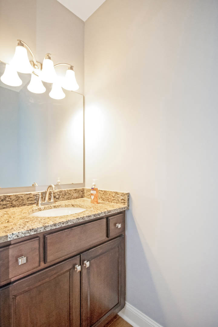 Marble countertop vanity with undermount sink, chrome faucet, brown cabinetry, wall-mounted mirror, and ceiling light fixture in a tiled bathroom.