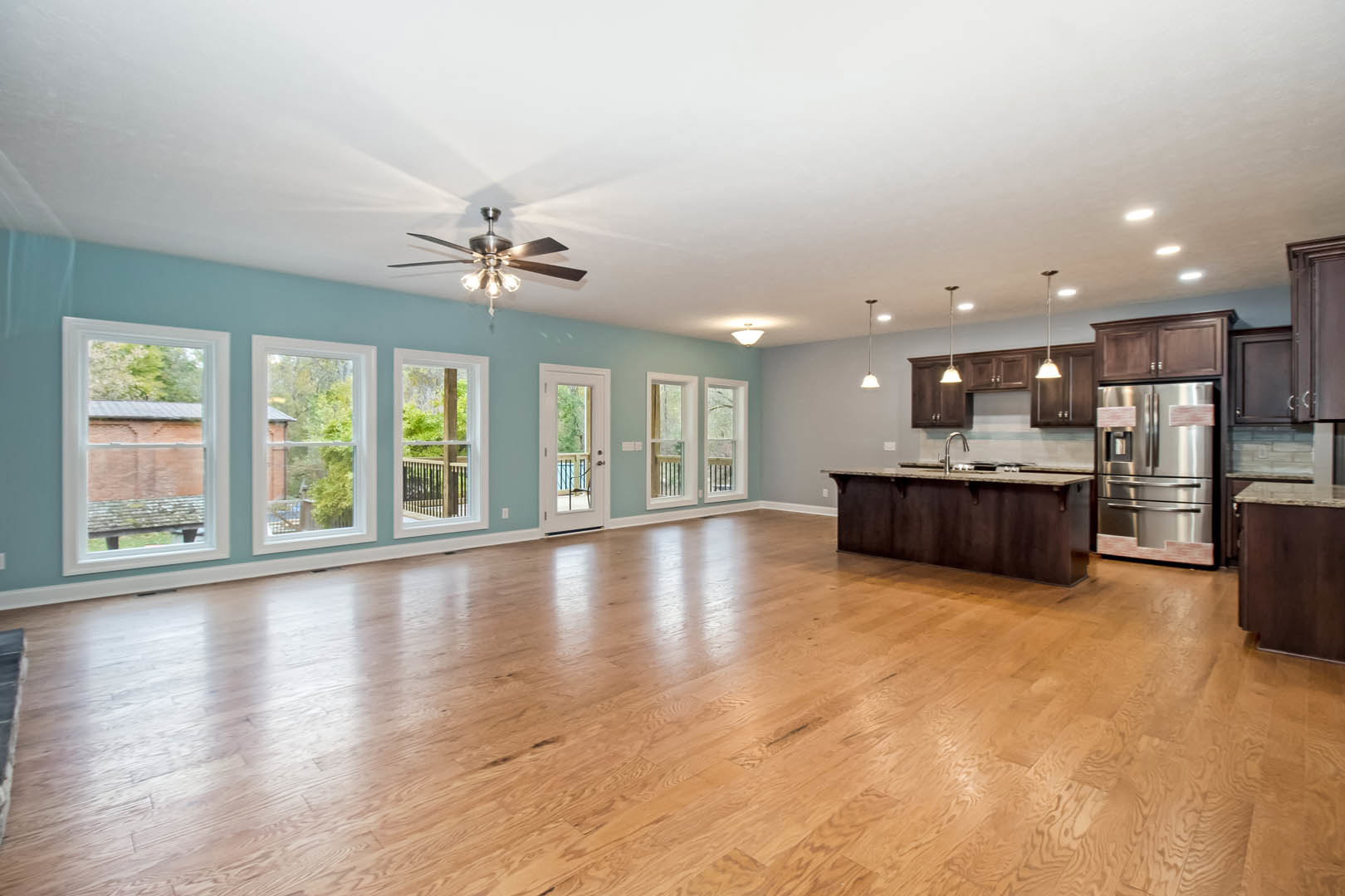 Open-concept kitchen with stainless steel refrigerator, hardwood floors, white cabinetry, large windows overlooking brick wall and trees, and glass door leading to balcony