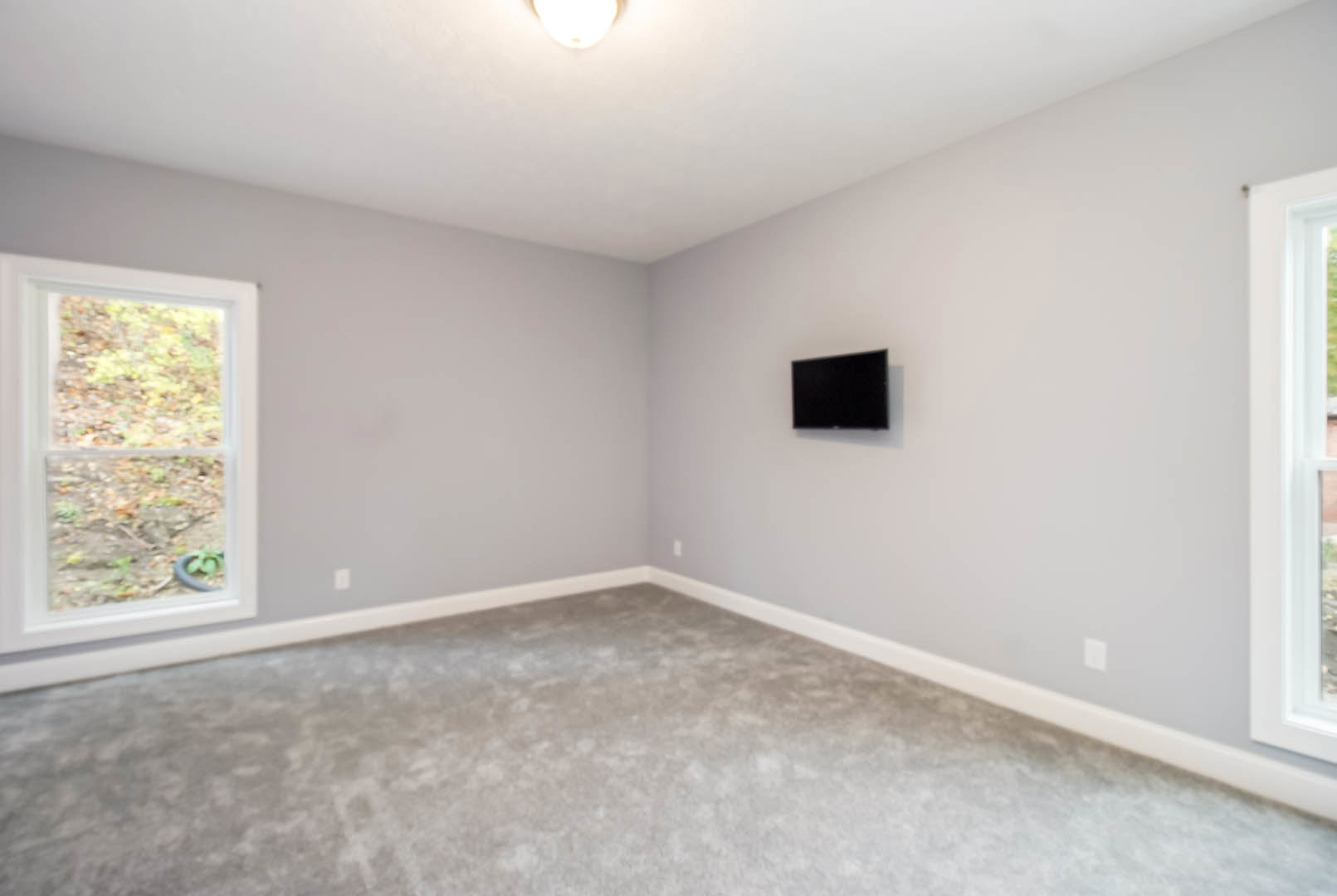 Television mounted on a white wall above grey carpeted floor, large window with view of dirt and plants, simple molding and plaster finishes