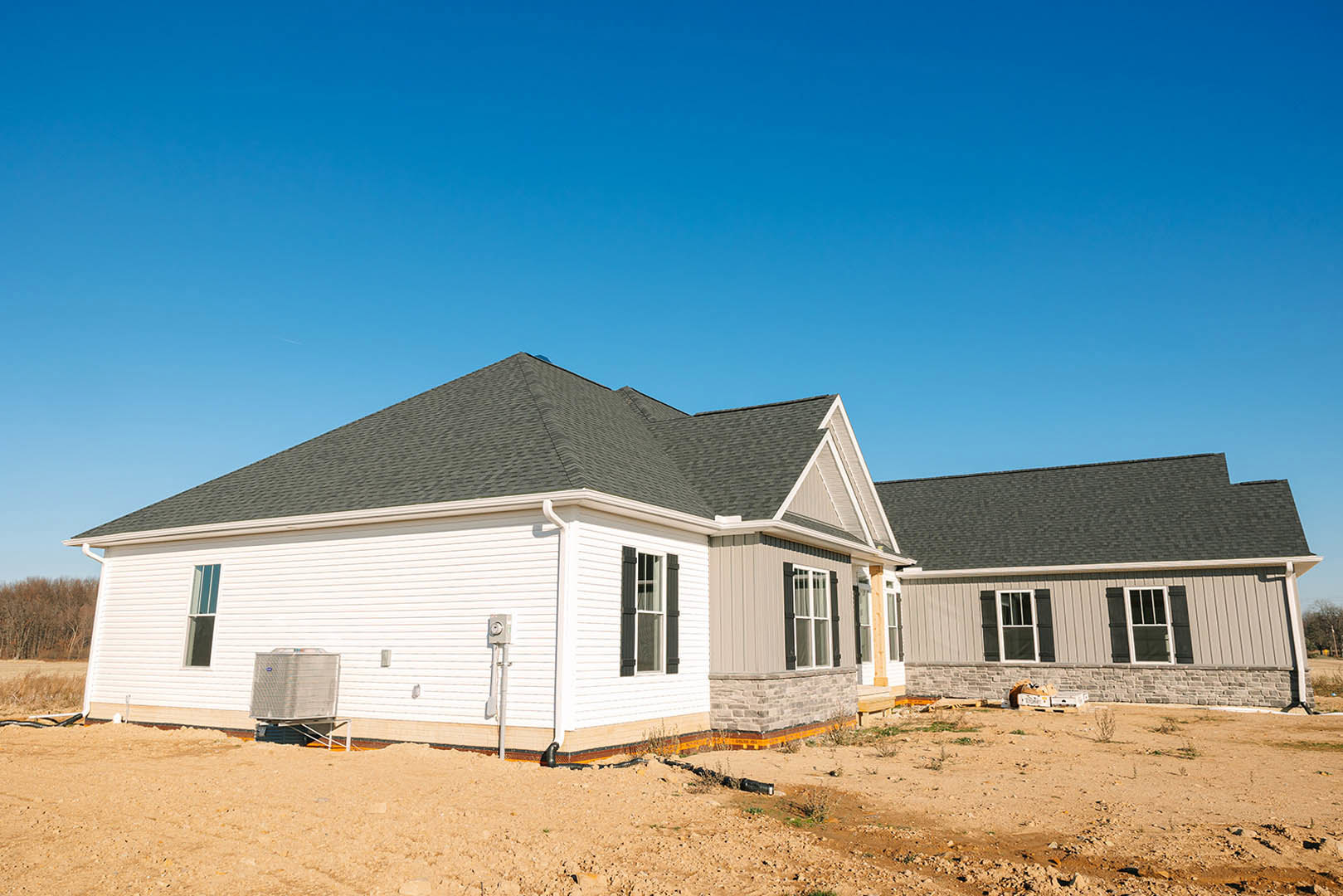 Framed house under construction with exposed roof trusses, white window frames, white siding, dirt foreground, large metal container on trailer, blue sky, and neighboring homes in