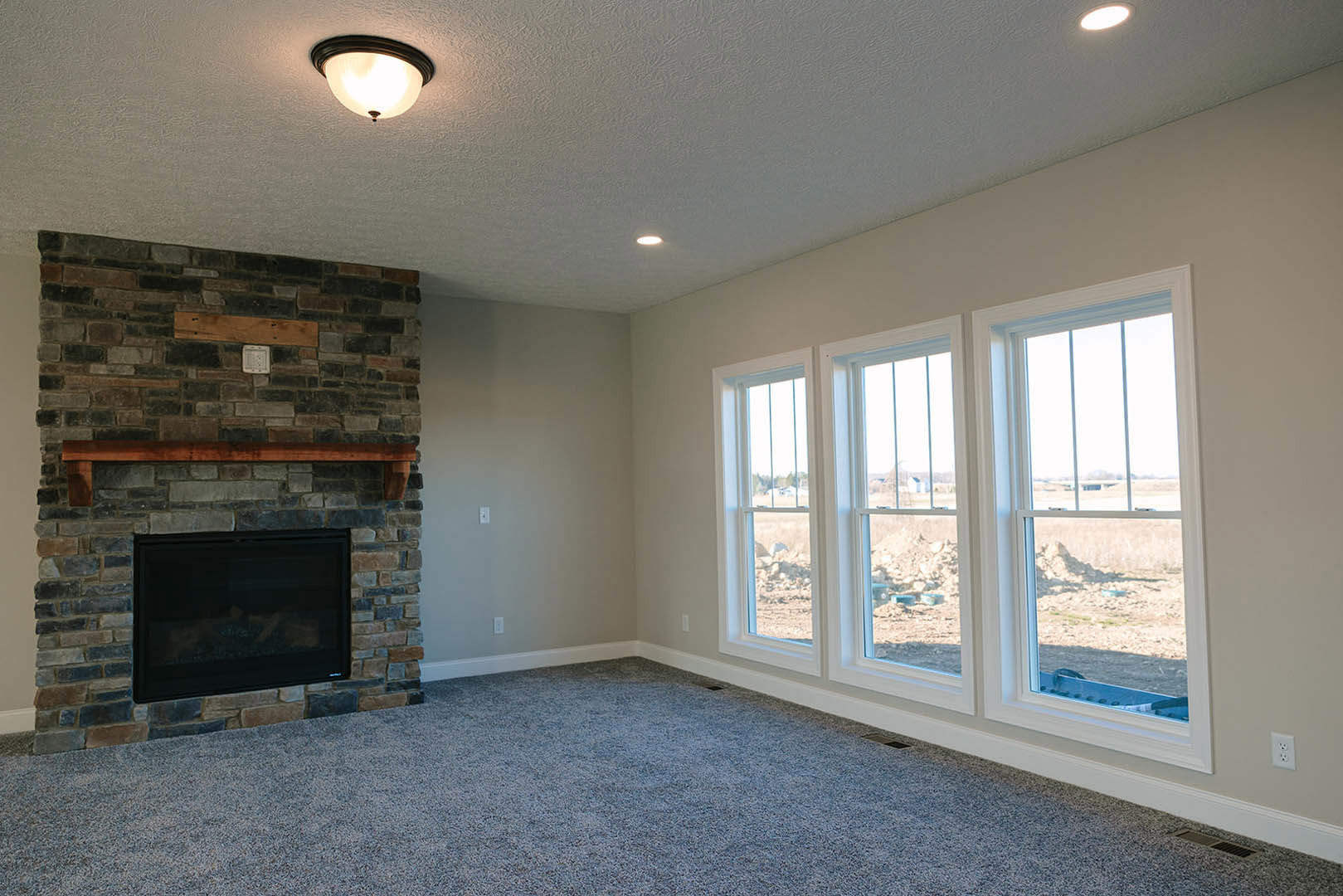 Carpeted living room featuring a stone fireplace with wooden mantel, large windows framed by white trim, and recessed ceiling lights
