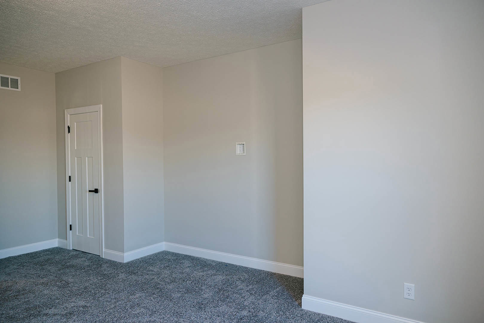 White carpeted floor, white wall with black baseboard, white door featuring black handle, electrical outlet visible on wall