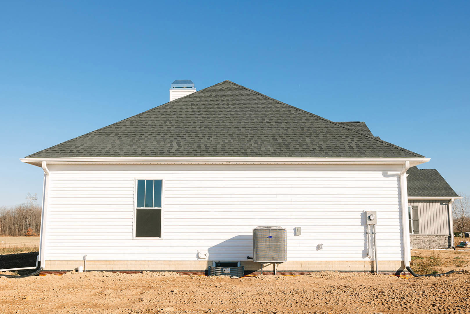 White house with black roof, horizontal siding, large windows, grassy yard, and metal barrel on dirt lot