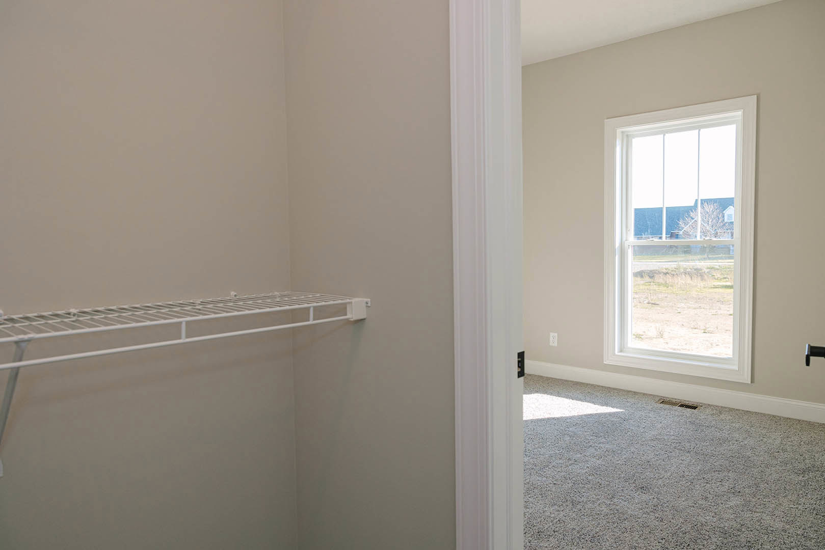 Carpeted room with white metal shelf against white wall, window overlooking tree and neighboring house, floor vent visible