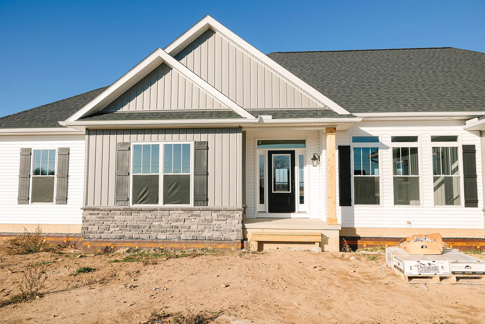 Modern house under construction with white siding, black door with glass panes, stone accent wall, large window, and dirt yard with cardboard box on pallet
