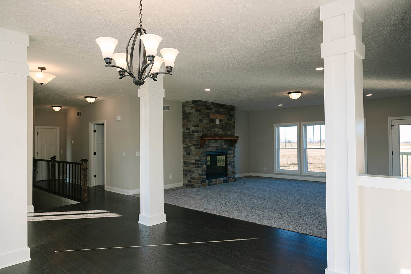 Spacious living room featuring a stone fireplace with wood mantel, elegant chandelier hanging from plaster ceiling, large window set in brick wall, black metal railing, and white