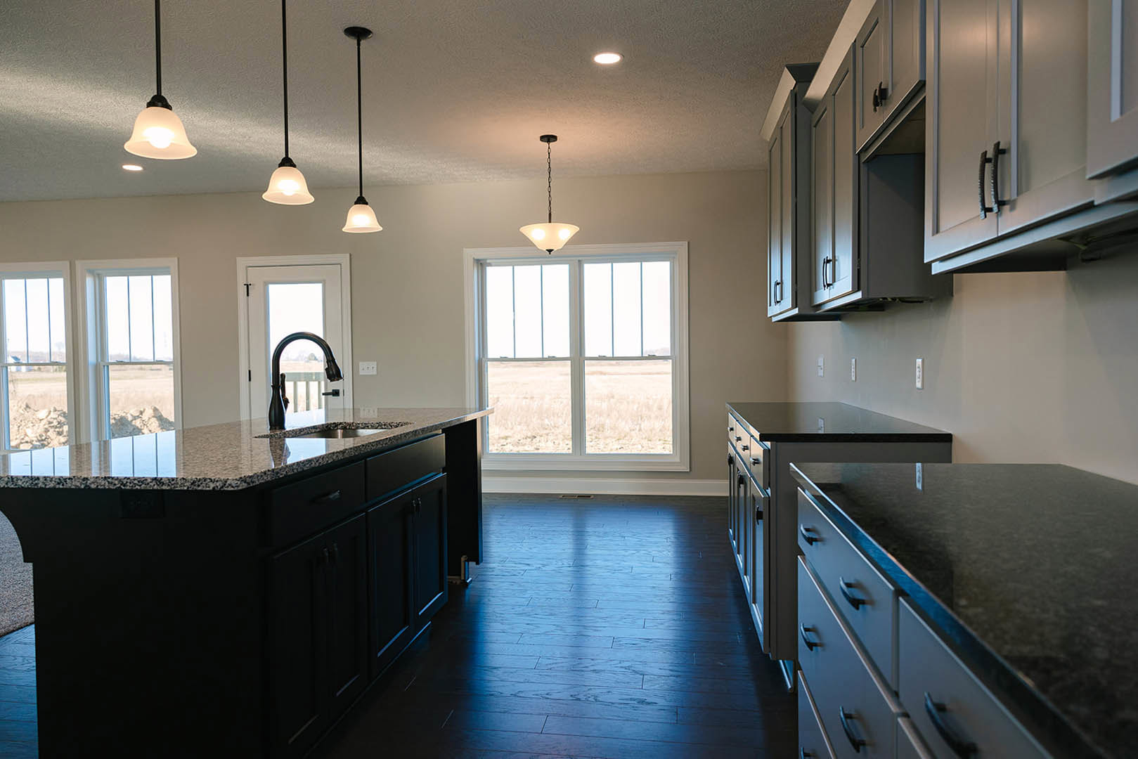 Spacious kitchen featuring a large island with built-in sink and black faucet, light wood cabinetry with drawers, stone countertops, pendant light fixture near window, white walls