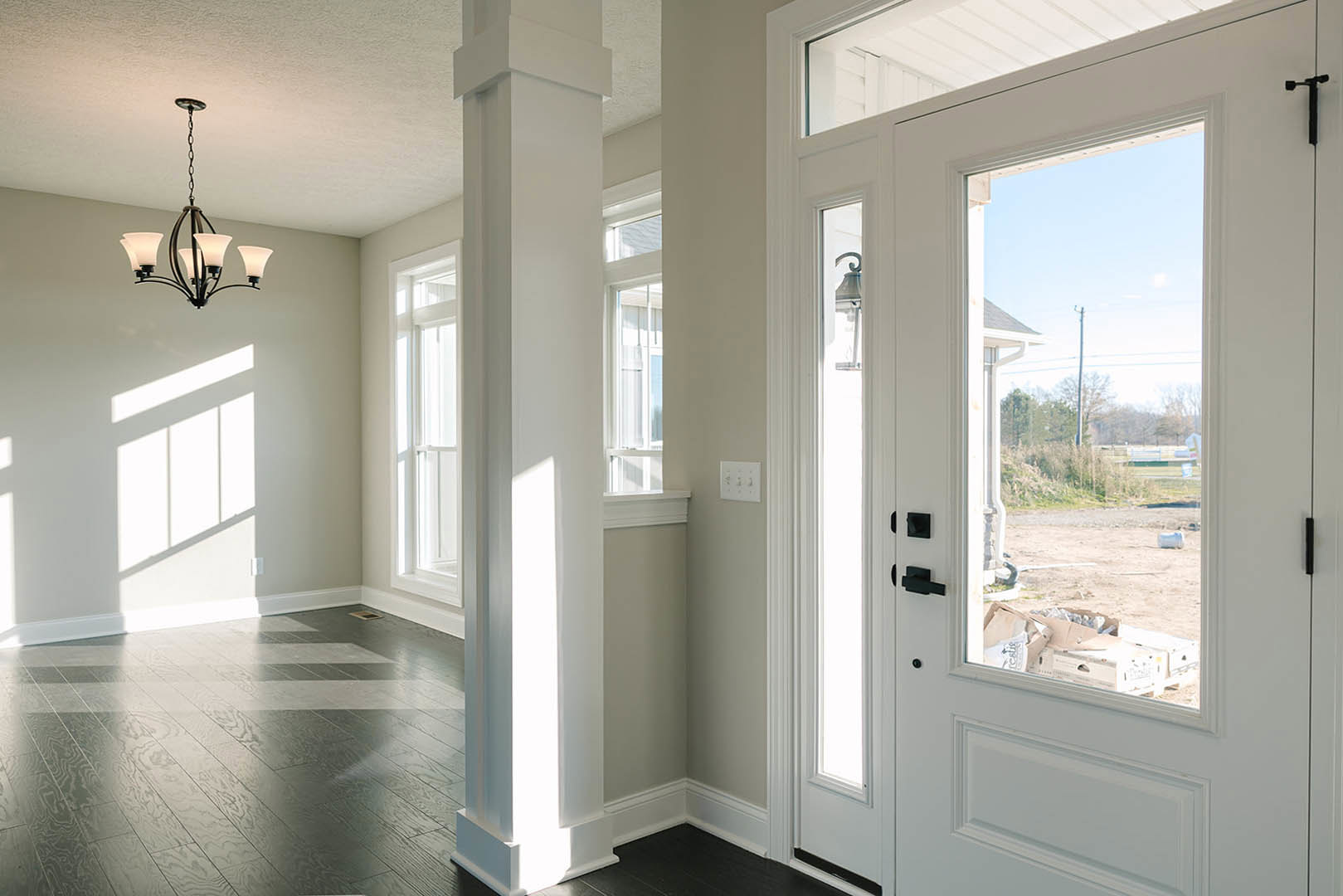 White paneled door beside a white column, dark wood flooring with white trim, chandelier with white shades, white wall with light switch, lamp close up, natural light filtering