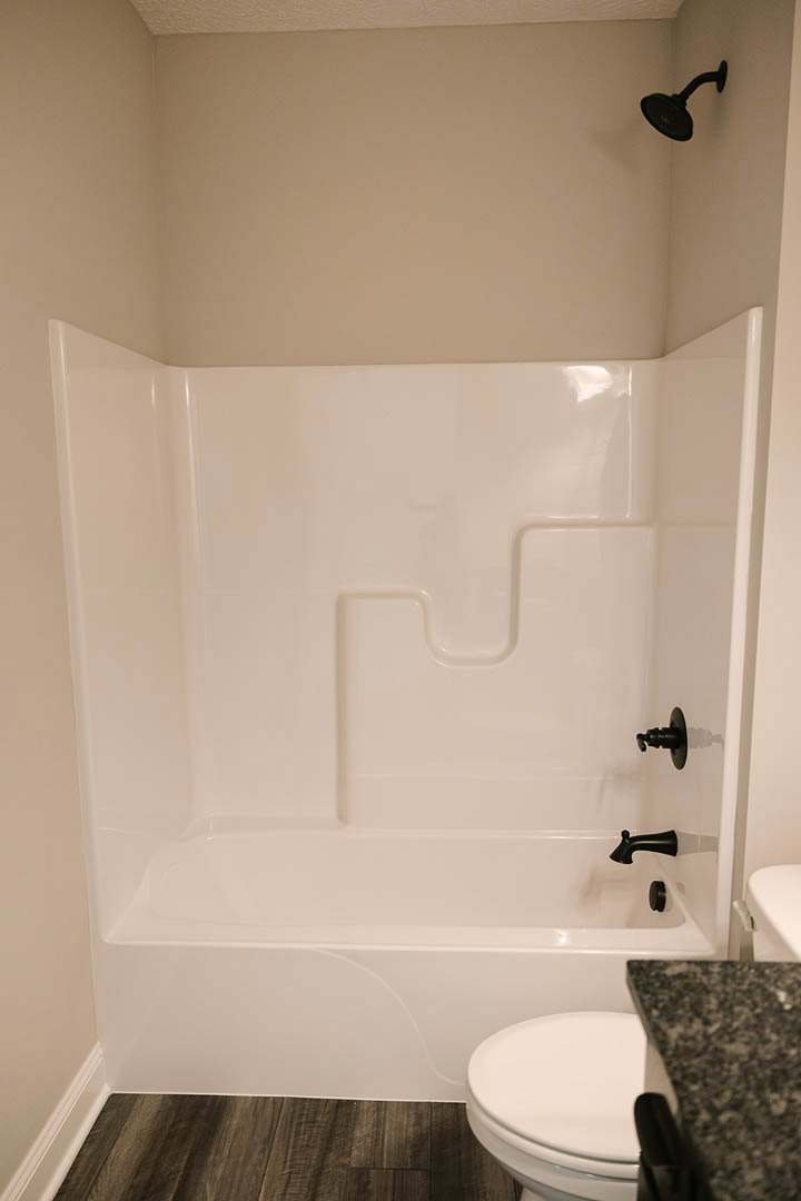 Freestanding white bathtub beside white toilet, wood vanity with stone countertop, tiled walls, chrome shower head