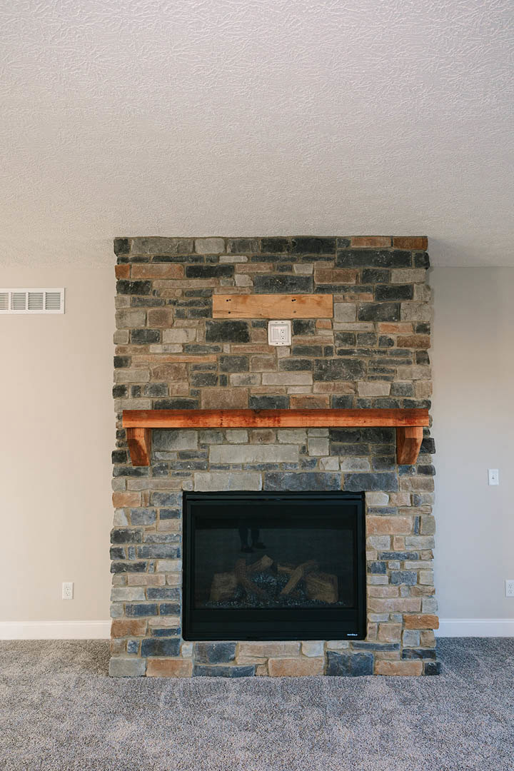 Brick fireplace with wood mantel, stacked logs in hearth, stone hearth, fire screen, carpeted floor, wood beam accent, indoor living room