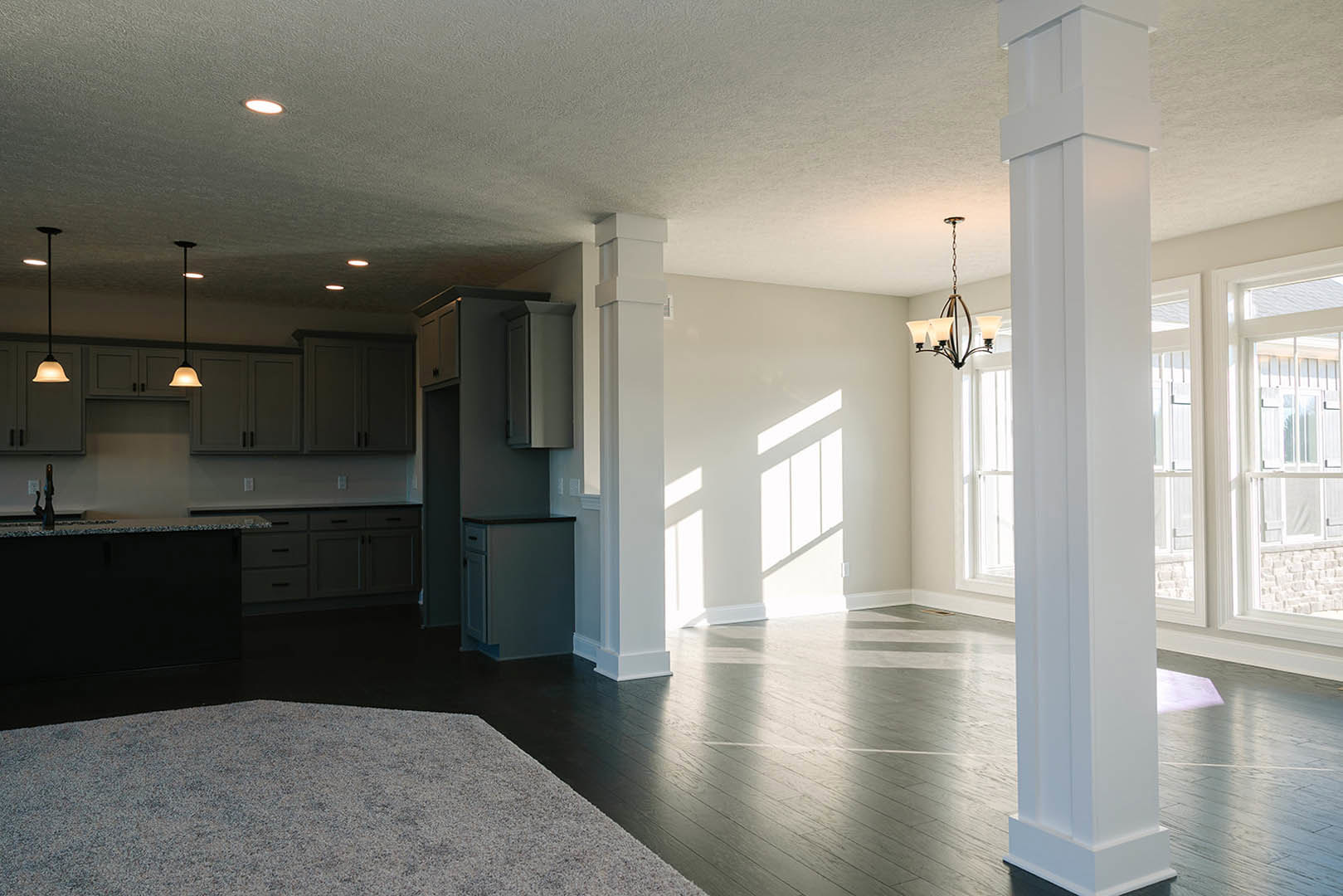 White walls and dark hardwood flooring in a spacious room, featuring a white pillar, modern chandelier, and black rectangular accent with blue surface; white square with black trim