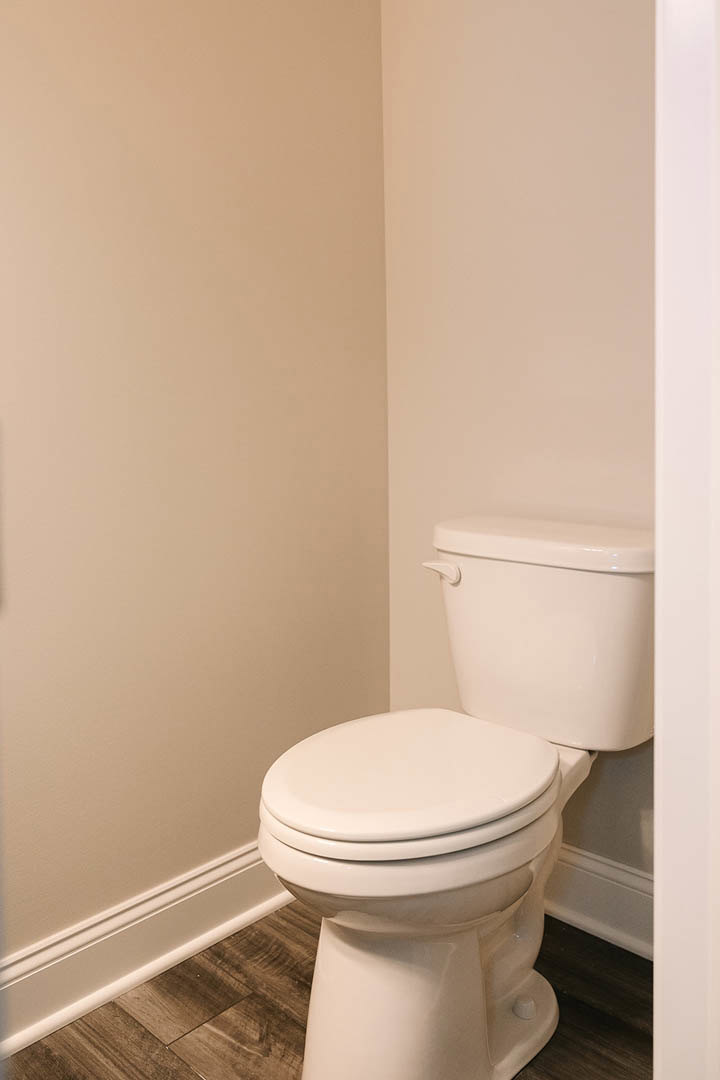 White toilet with closed lid positioned in a corner of a small bathroom, surrounded by light-colored tile walls and wood flooring
