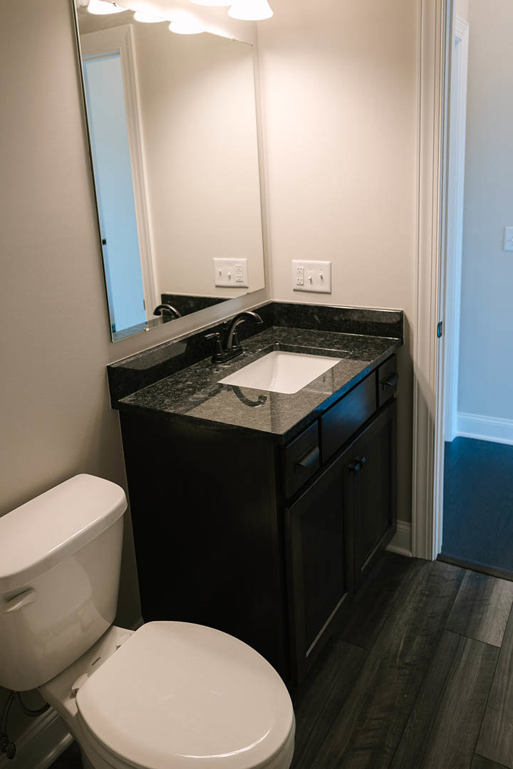 White toilet beside black marble countertop with undermount sink, wall-mounted mirror, and light switch on tiled bathroom wall