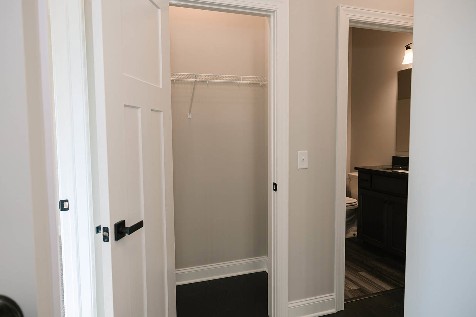 Open closet door revealing white walls, dark wood flooring with white base trim, adjacent dark cabinet and toilet, close-up of silver door handle, mirror reflecting bathroom