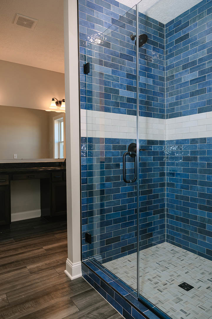 Blue tile shower with glass door, tile wall featuring shower head, ceiling vent, and wood flooring adjacent to blue tile.