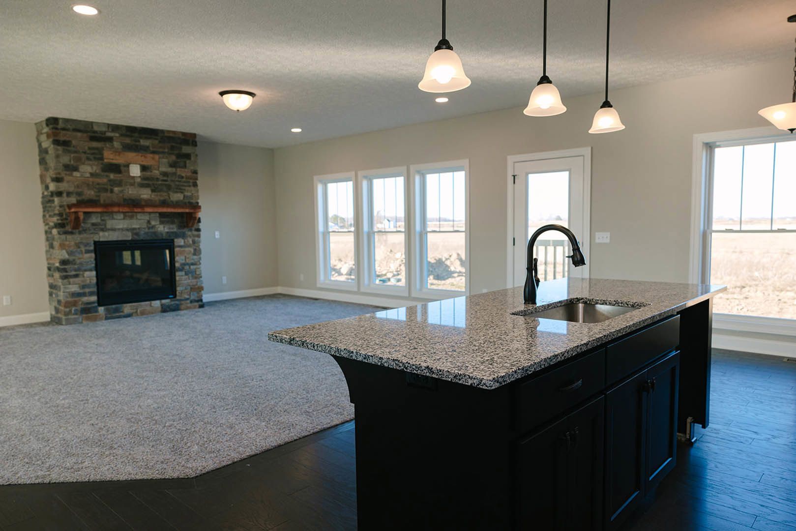 White kitchen island with built-in sink and chrome faucet, black rectangular range hood on exposed brick wall, fireplace below mounted TV, row of windows overlooking grassy field