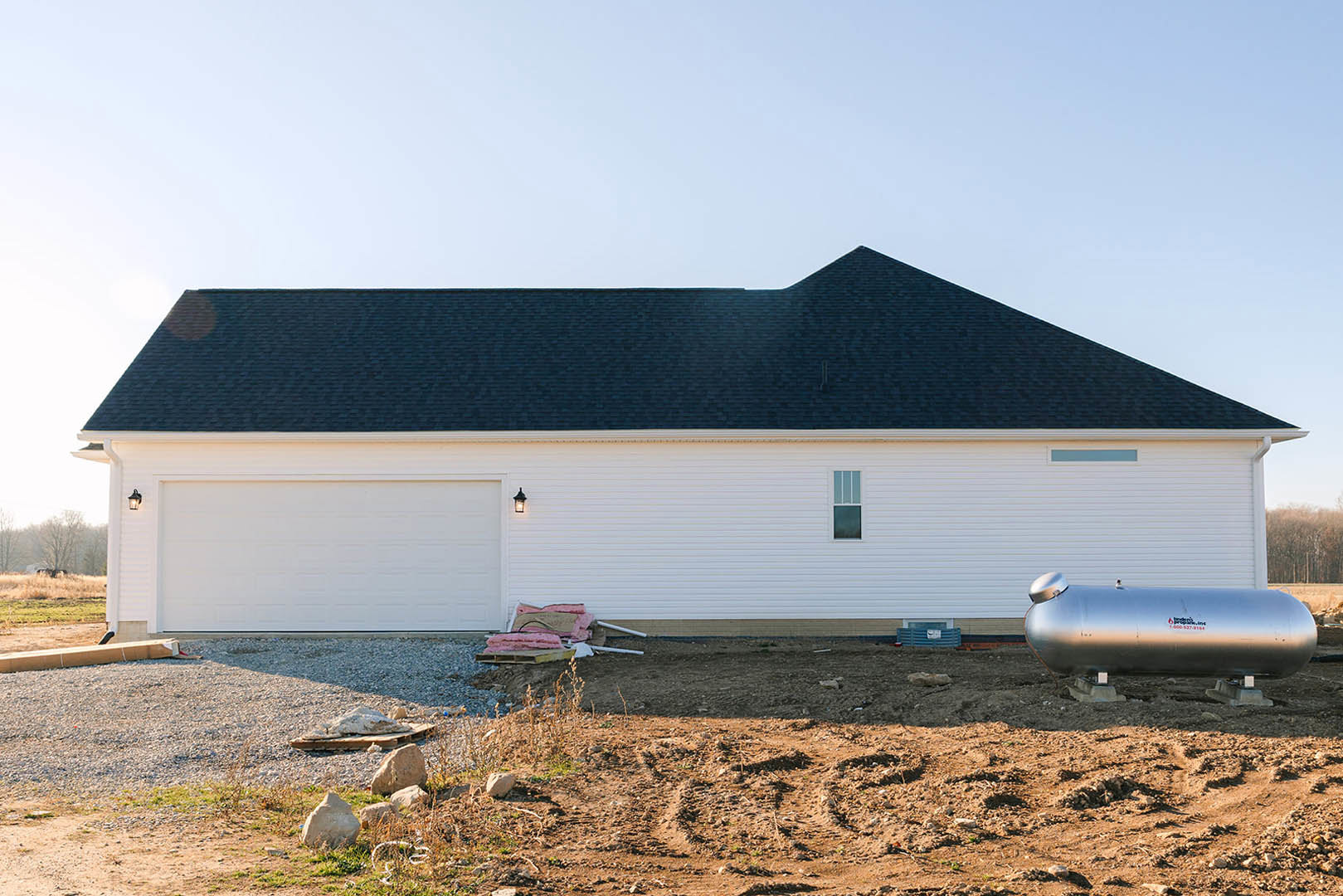White house with black roof, attached garage, large water tank beside building, grassy yard with scattered rocks.