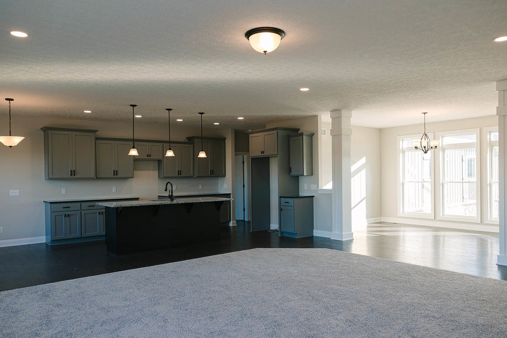 Open-concept kitchen and living room featuring a large patterned area rug, black countertop with matching cabinetry, white door, tile flooring, and ceiling chandelier.