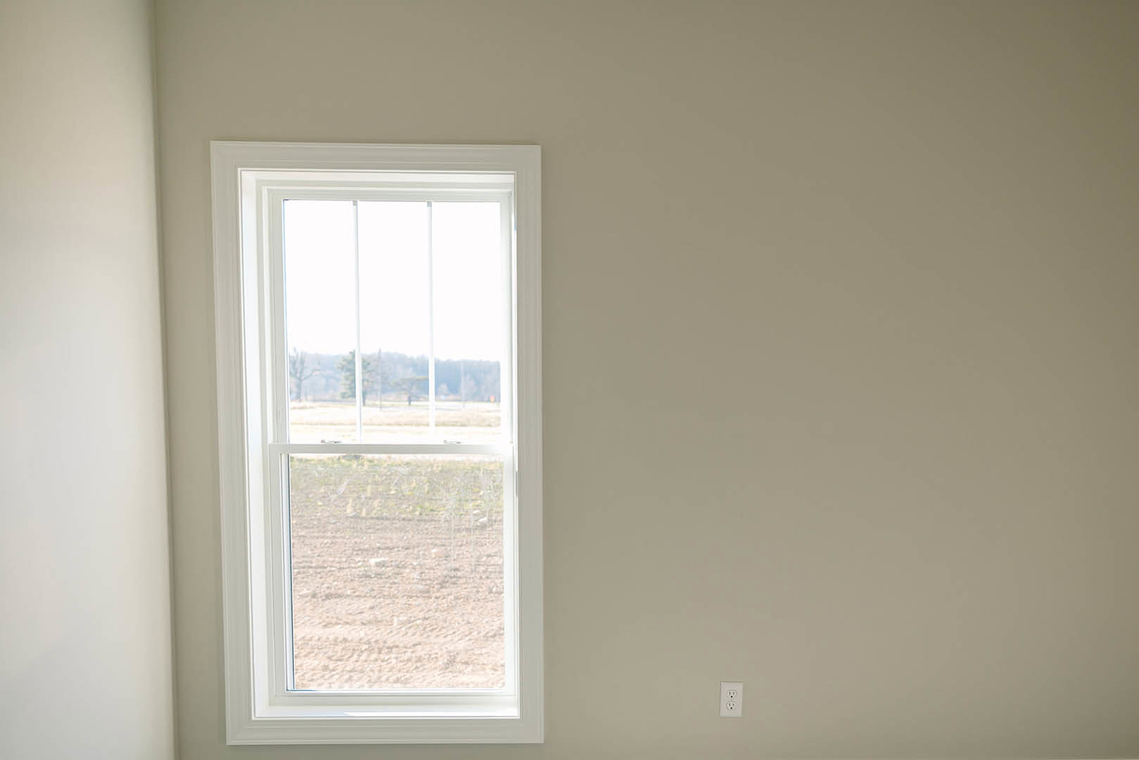 White plaster wall with large window, sheer blinds partially drawn, daylight streaming in, view of grassy field and distant trees outside