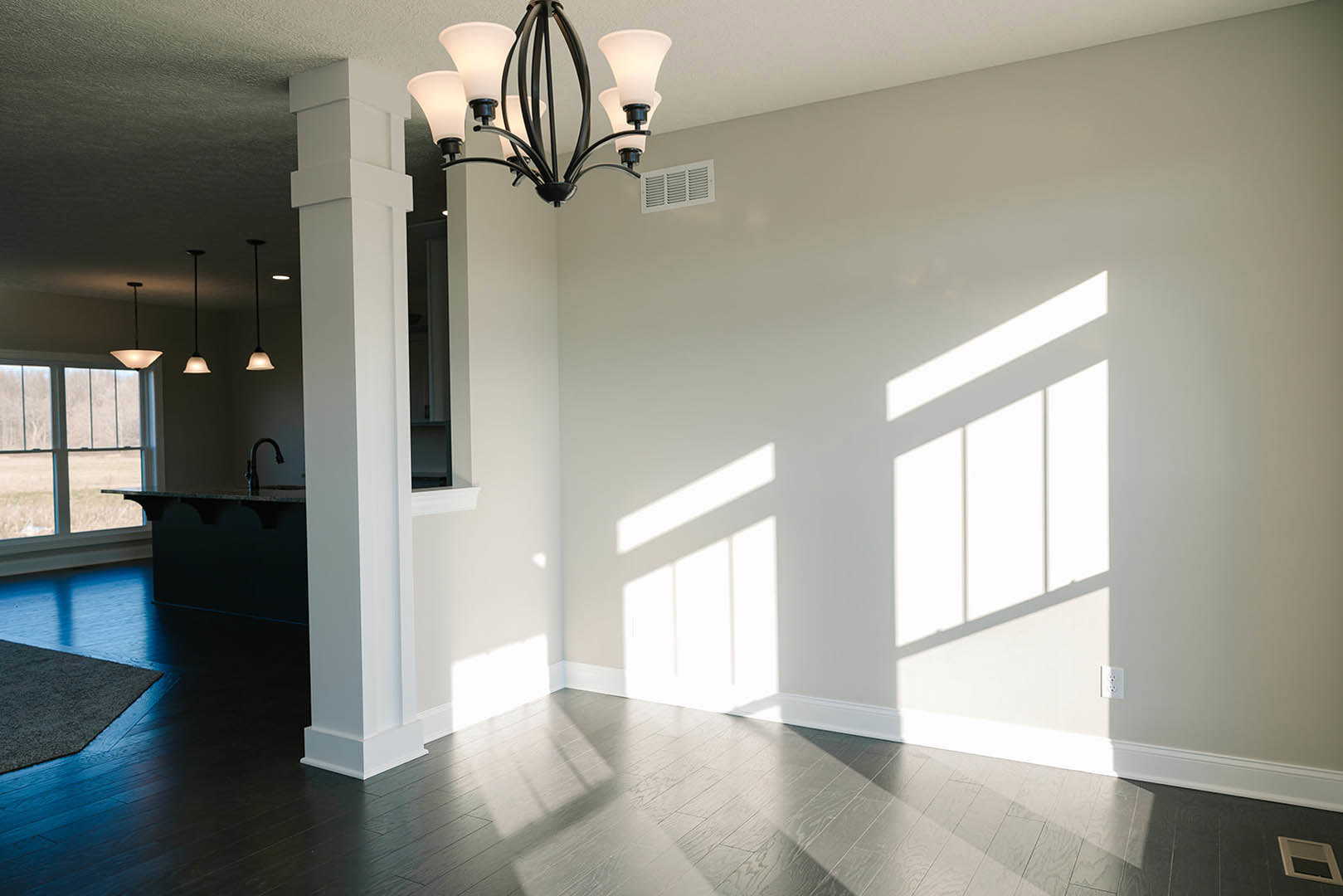 Chandelier hanging from white plaster ceiling, wood flooring, wall vent, partial view of area rug and chrome faucet, natural daylight illuminating interior space