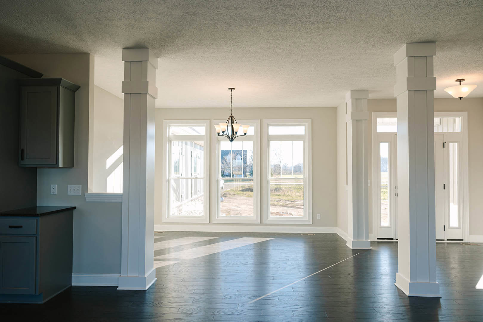 Open living space with white pillars, ornate chandelier, wood flooring, white paneled door, built-in cabinetry, and decorative molding