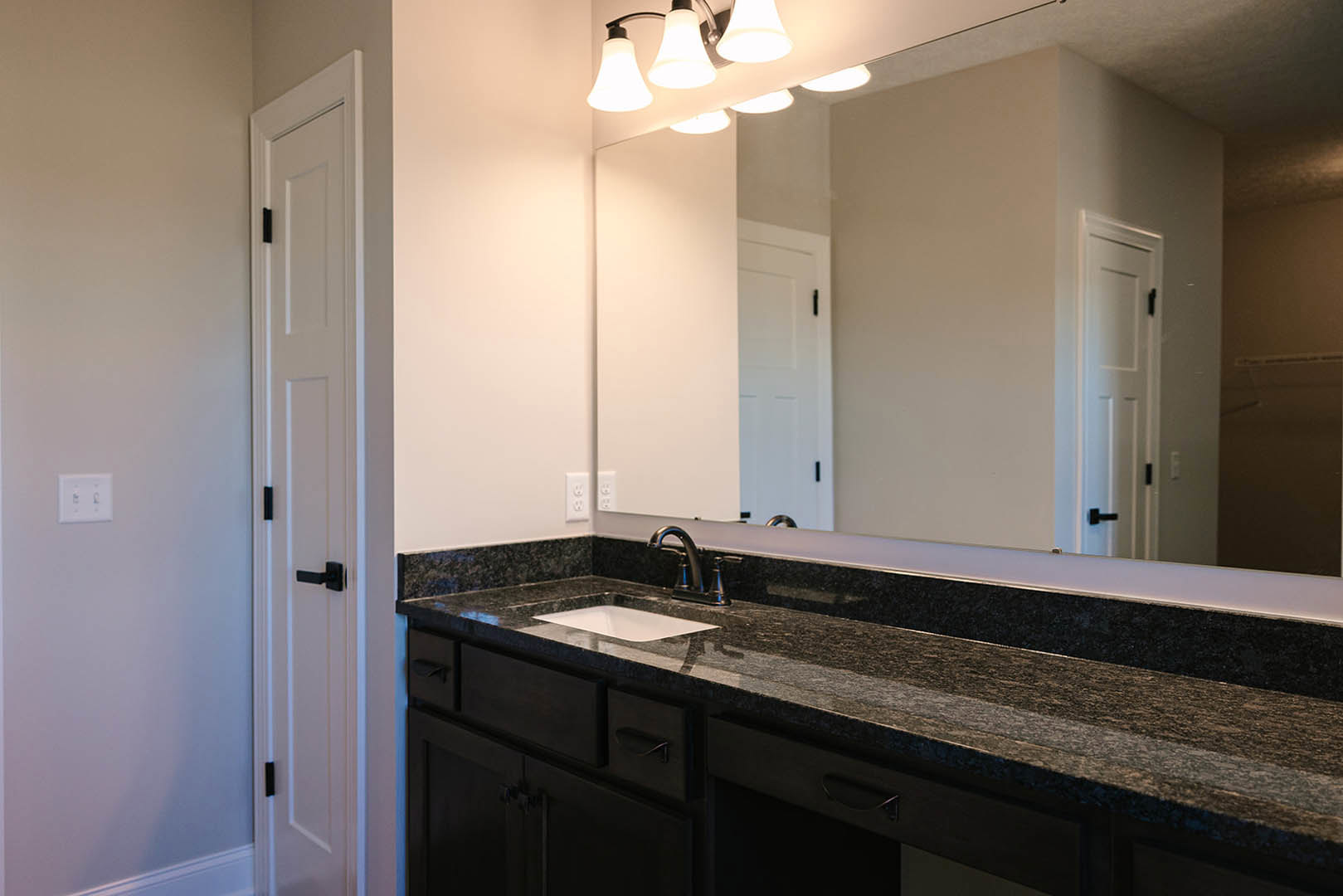 Black granite countertop with black faucet and white sink beneath a wall-mounted mirror, white cabinetry, and tiled backsplash
