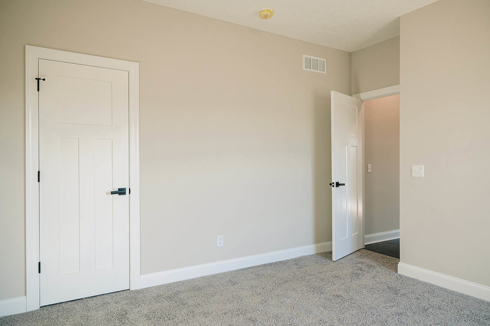 Bedroom with plush beige carpet, two white paneled doors featuring black handles, white walls, and a wall-mounted vent