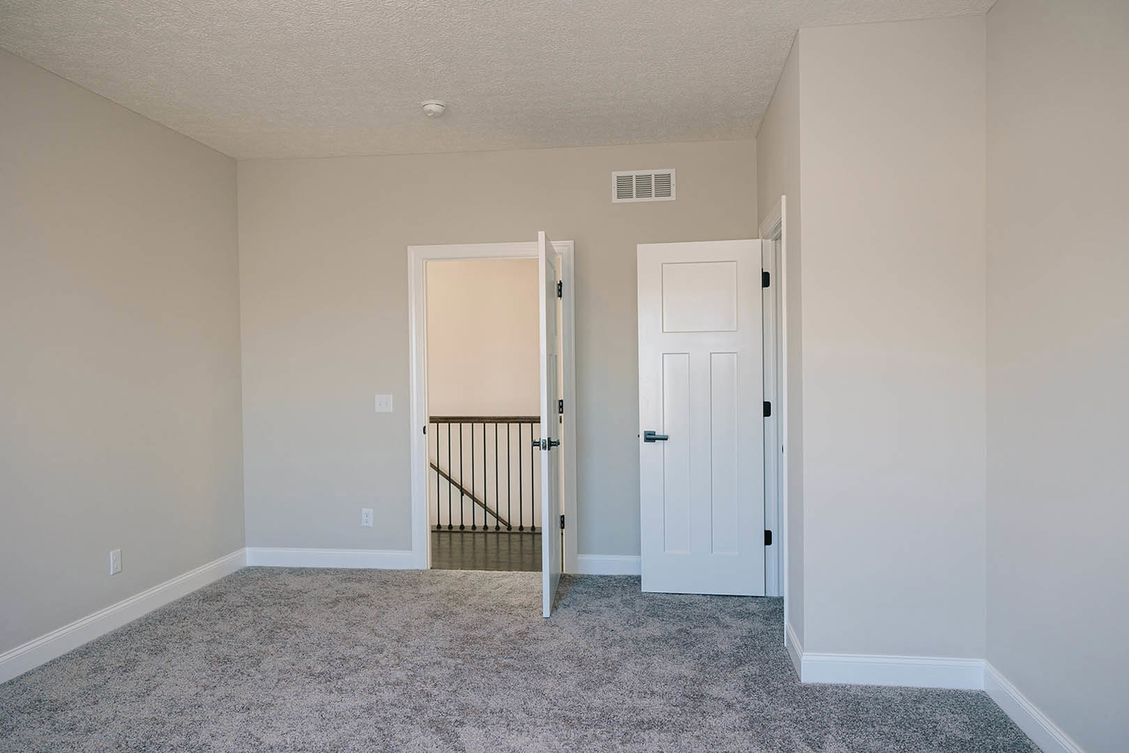 Carpeted walls and floor in a room with two doors, one white with a black handle and one open to a staircase, wall vent and metal railing visible