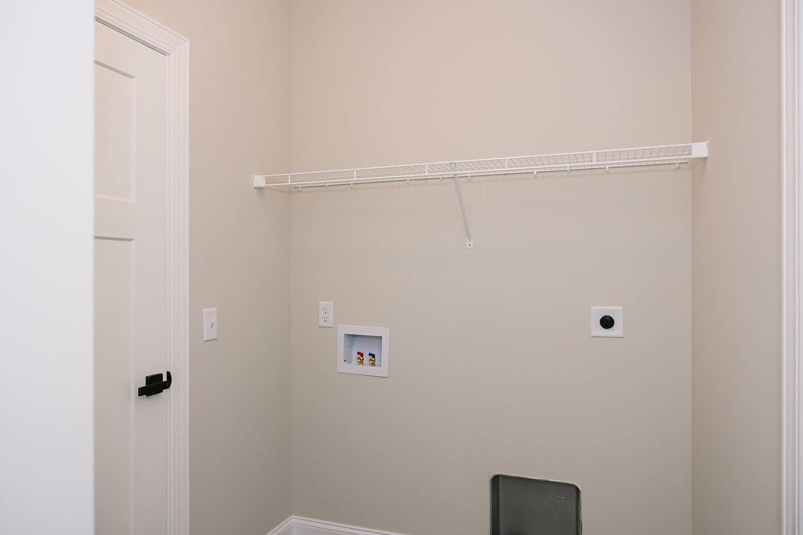 White shelf mounted on smooth plaster wall beside white door with black handle, adjacent to grey utility box and round black fixture, bathroom interior with neutral finishes.