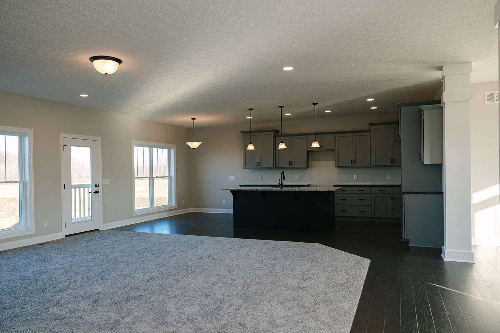 Spacious open-concept interior featuring a kitchen island with tile flooring, cabinetry, ceiling fixtures, a patterned rug, white-framed windows, and a white door with glass panel.