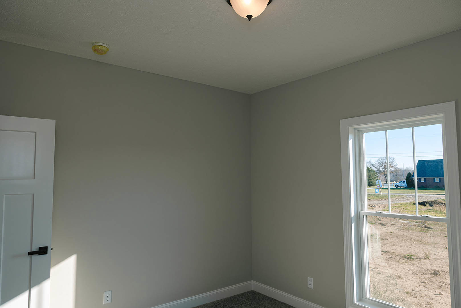 White door and trim, yellow ceiling light fixture, window overlooking grassy field and parked truck, neutral plaster walls, light wood floor.